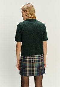 Dunkelgrüner kurzärmeliger Pullover mit geripptem Saum, kombiniert mit einem Karorock in den Farben Marineblau, Grün und Beige. Geschmeidige Textur.