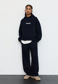 Kobieta w granatowej bluzie z kapturem z napisem "New Studio Rotterdam", dopasowanych szerokich spodniach, białej koszuli pod spodem i beżowych trampkach stojąca na tle białej ściany.