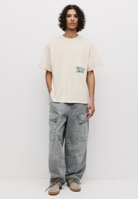 Beige bomulds t-shirt med en brystlomme og grøn grafisk tekst. Sammen med løstsiddende, grå denim cargo bukser og lysebrune sko.