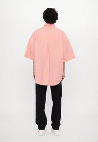 Camisa de gran tamaño de color rosa claro con mangas cortas, detalle en el yugo posterior, dobladillo curvado, combinada con pantalones negros y zapatos blancos. Textura lisa.