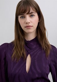 Vestido morado con cuello alto, que presenta un corte en forma de gota y un patrón texturizado. Confeccionado en tela suave, con detalles de mangas abullonadas en los hombros.