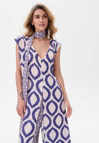 Fioletowy i kremowy kombinezon w wzory z głębokim dekoltem w kształcie V, krótkimi rękawami oraz pasującym szalem związanym na szyi. Gładka tekstura materiału.