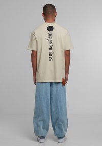 Béžové bavlněné tričko s černým textem na zadní straně, který zní "zapomenuté zákony", v kombinaci se světle modrými širokými džínovými kalhotami a bílými teniskami.