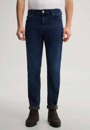 Mann trägt dunkelblaue hochgekrempelte Jeans, weißes Hemd und dunkelbraune Wildlederstiefel und steht vor einem schlichten hellen Hintergrund.
