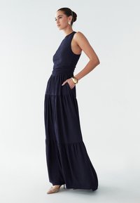 Tumšzils maksi kleita ar fakturētu audumu, pieguļošu bodiju, vairāksluoksnētu svārku un sānu kabatām. Piedāvā bezroku dizainu un gludu kakla izgriezumu.