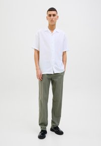 Joven de pie vistiendo una camisa blanca de manga corta con botones, pantalones verde oliva y zapatos negros, sobre un fondo claro y liso.