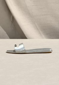 Φλατ ασημένιο σανδάλι με ανοιχτό δάχτυλο, διακοσμημένο με μια μικρή μεταλλική μπάλα, σε ουδέτερο μπεζ φόντο και επιφάνεια.