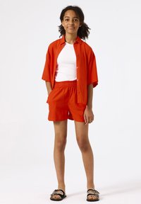 Camisa roja de manga corta sobre una camiseta blanca de tirantes, combinada con pantalones cortos rojos y sandalias negras con detalles blancos. Tejido suave y corte relajado.