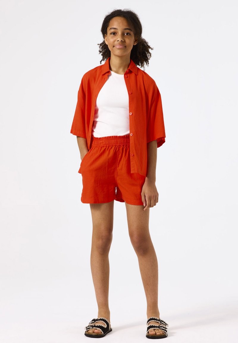 Camisa roja de manga corta sobre una camiseta blanca de tirantes, combinada con pantalones cortos rojos y sandalias negras con detalles blancos. Tejido suave y corte relajado.