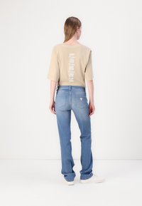 Beige oversized short-sleeve shirt with vertical printed text on the back, paired with light blue straight-leg denim jeans. White sneakers visible.