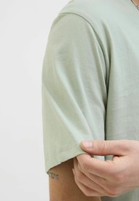 Světle zelené bavlněné tričko s krátkými rukávy, s jemnou texturou a mírně volným střihem. Spodek je jednoduchý s čistým šitím.