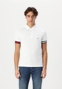 Hvit polo-skjorte med knappestolpe, har svarte og røde striper på ermene og en liten brodert logo på brystet.