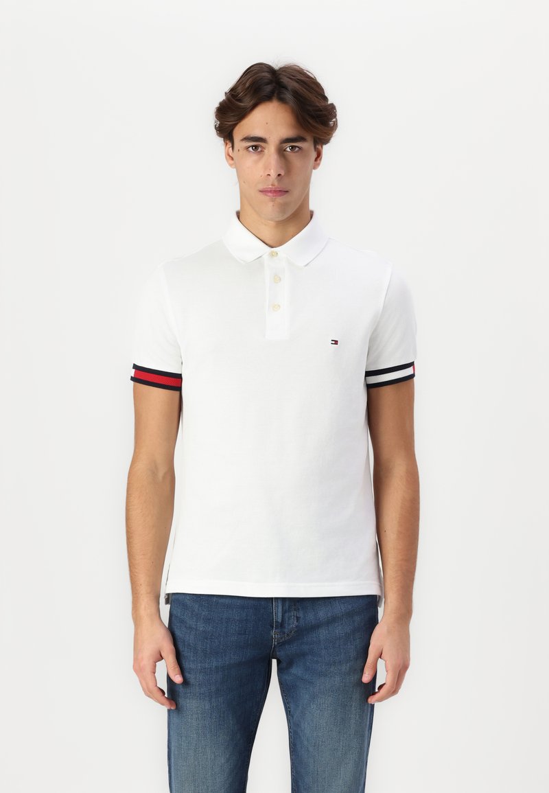 Hvit polo-skjorte med knappestolpe, har svarte og røde striper på ermene og en liten brodert logo på brystet.
