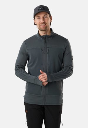 Mand iført en mørkegrå lynlåstrøje med tommelhuller og en sort kasket, stående foran en ensfarvet lys baggrund.