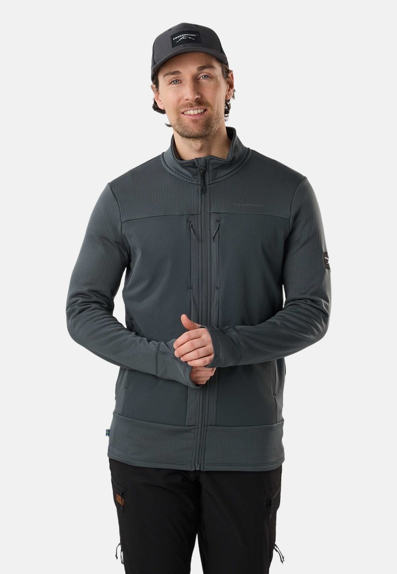 Homme portant une veste zippée gris foncé avec des ouvertures pour les pouces et une casquette noire, debout devant un fond clair uni.