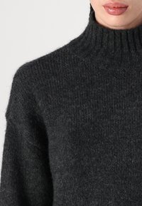 Gros plan sur une personne portant un pull à col roulé en tricot côtelé gris foncé, montrant le col et la partie supérieure de la poitrine.