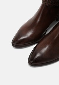 Botas de cuero marrón hasta el tobillo con un acabado suave y brillante, punteras puntiagudas y un detalle de textura tejida en la parte superior. Suelas planas.