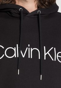Czarna bluza z kapturem z białym logo "Calvin Klein", posiadająca kieszeń typu kangur, sznurki ze metalowymi końcówkami oraz prążkowaną teksturę.