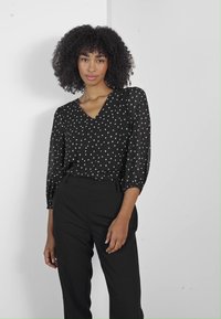 Blouse noire à pois blancs, décolleté en V, et manches trois-quarts. Associée à un pantalon noir taille haute. Texture lisse.