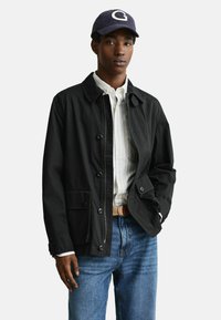 Junger Mann mit einer schwarzen, geschlossenen Jacke, einem weiß gestreiften Hemd, einer blauen Jeans, einem beigen Gürtel und einer marineblauen Kappe mit einem weißen "G" auf der Vorderseite.