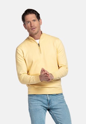 Man in een gele trui met kwart rits en lichtblauwe spijkerbroek die zijn handen in elkaar vouwt, staand tegen een effen lichte achtergrond.