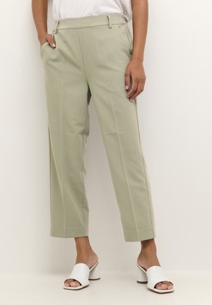 Pantaloni verde chiaro con taglio sartoriale, dotati di tasche laterali e una texture liscia. Abbinati a scarpe con tacco aperte bianche.