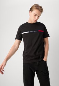 Schwarzes Baumwoll-T-Shirt mit einem Logo auf der Front und horizontalen Streifen in Weiß und Rot. Klassischer Rundhalsausschnitt und kurze Ärmel.