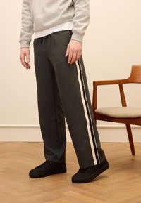 Grijze joggings met crèmekleurige zijkanten, gemaakt van een gladde stof. Gecombineerd met zwarte sneakers, tegen een lichte achtergrond en een houten stoel.