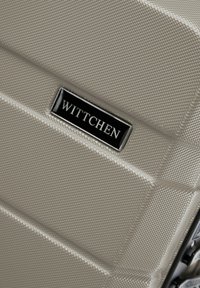 Valigia rigida beige con finitura strutturata. Presenta accessori neri e un logo argentato "Wittchen" su una placca rettangolare.