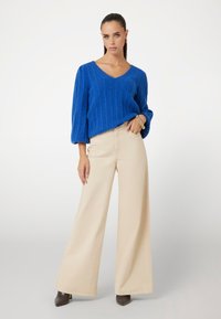 Maglione blu a coste con scollatura a V e maniche larghe e corte, abbinato a pantaloni beige a gamba larga e scarpe con tacco marroni.