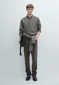 Skjorta med knappar i grön och svart rutigt mönster, grå tröja runt midjan, grå byxor och svarta skor. Modellen står mot en ljus bakgrund.