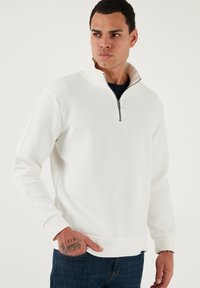 Biely žebrovaný pulóver s golierom na štvrtinový zips, dlhými rukávmi a pevným lemom, kombinovaný s modrými džínami. Viditeľný detail textúrovaného materiálu.