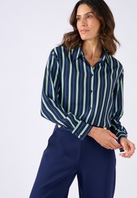 Chemise rayée bleu marine et verte à manches longues, avec un col à boutons et des poignets boutonnés, assortie à un pantalon bleu marine ajusté. Texture de tissu lisse.
