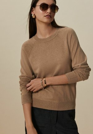 Beige gestrickter Pullover mit Raglanärmeln, Rundhalsausschnitt und gerippten Bündchen. Modell trägt große Sonnenbrillen und goldene Accessoires. Dunkle Hose.