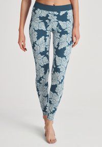 Femme portant des leggings bleus avec de grands motifs floraux blancs, se tenant pieds nus devant un fond neutre.