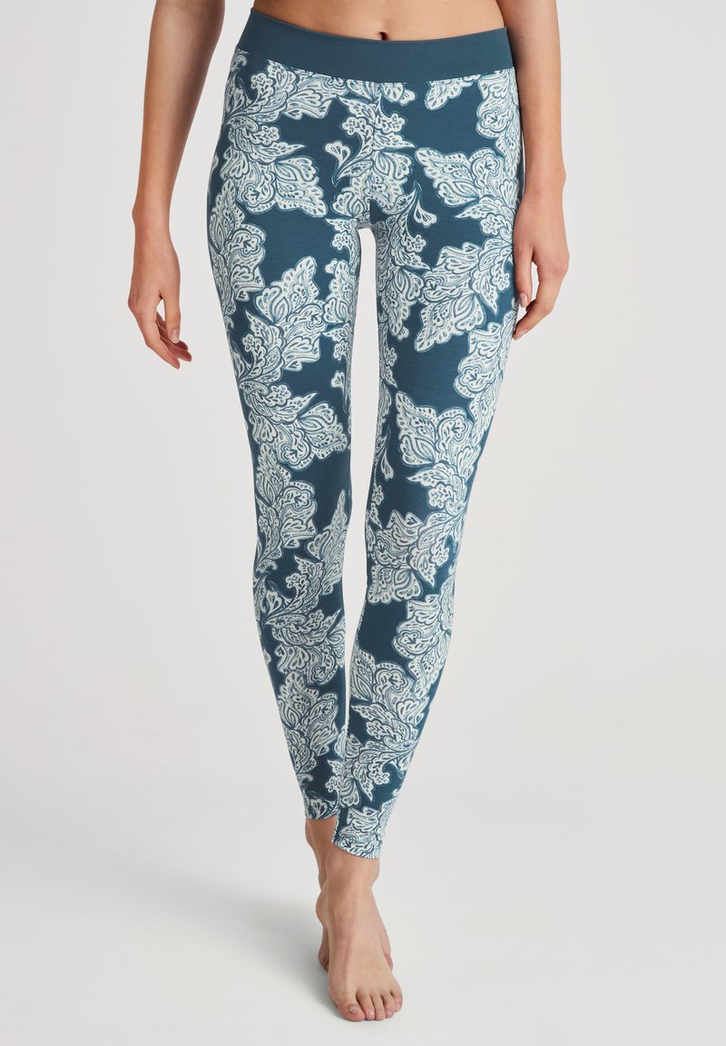 Femme portant des leggings bleus avec de grands motifs floraux blancs, se tenant pieds nus devant un fond neutre.