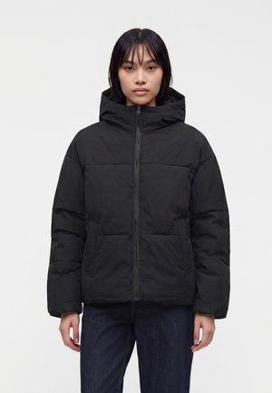 Persona joven con cabello oscuro y lacio que lleva una chaqueta acolchada negra con capucha y cremallera frontal, posando frente a un fondo blanco liso.