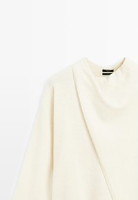 Pull crème avec un col drapé et une texture douce, présentant une coupe décontractée et des épaules tombantes. Étiquette visible au niveau du col.