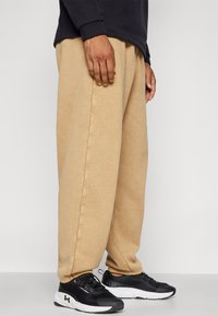 Beige, comfortabele broek van zacht materiaal met uitgesproken zijnaden. Gepaard met zwarte sportschoenen met witte accenten.