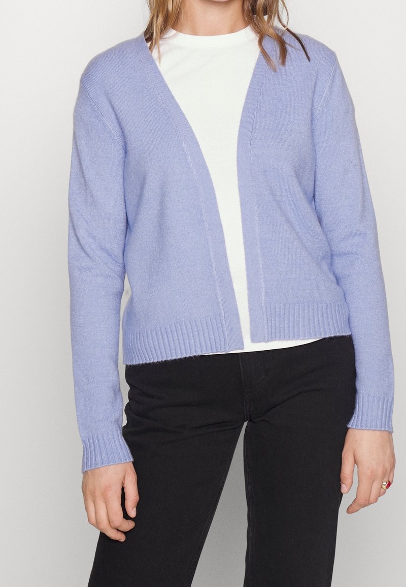 Cárdigan de punto en color púrpura claro con escote en V, puños y dobladillo acanalados. Se lleva sobre una camiseta blanca, presenta un ajuste holgado y una textura suave.