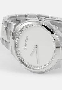Reloj de metal plateado con esfera blanca redonda, con manecillas de hora plateadas minimalistas, y un bisel adornado con pequeños cristales transparentes.