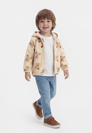 Lächendes Kind, das geht, trägt eine beige Fleecejacke mit Teddybär-Motiv, ein weißes Hemd, hellblaue Jeans und braune Turnschuhe vor weißem Hintergrund.