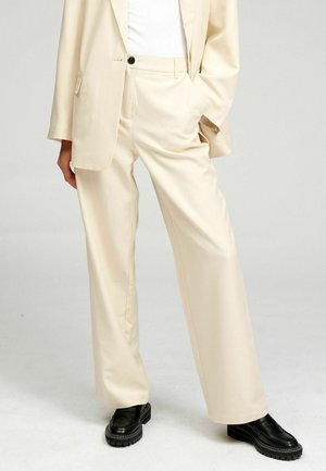 Krém színű, bőszárú nadrágot, hozzá illő blézert, fehér felsőt és fekete, vastag talpú mokaszinokat viselő személy, aki egyik kezét zsebében tartva áll.