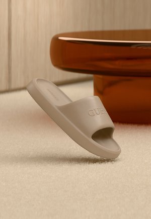 Sandalo beige slip-on con punta aperta e la scritta "GUESS" in rilievo sulla fascia, appoggiato su un tappeto chiaro vicino a un tavolo rotondo marrone.