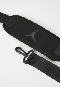 Melna neilona siksna ar gludu struktūru, kurai ir izdrukāts Jumpman logo, plastmasas klips, un redzamās šuvju detaļas.