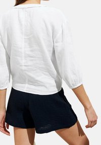 Blusa bianca in lino con maniche a sbuffo a tre quarti e scollatura rotonda, abbinata a pantaloni corti neri testurizzati. Un outfit semplice e casual.