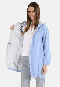 Veste imperméable bleu clair avec capuche, présentant un emblème circulaire rouge, des poignets élastiques et une fermeture éclair ; intérieur gris clair.