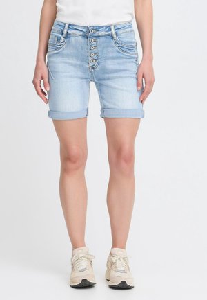 Femme portant un short en jean bleu clair retroussé avec quatre boutons sur le devant, des baskets blanches et un haut blanc sur un fond uni.