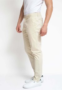 Chinos beige hechos de algodón con un corte ajustado. Cuenta con bolsillos delanteros, un bolsillo trasero y cierre de botón. Llevados con zapatillas blancas.