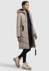 Beige Daunenjacke mit Kapuze, mit gestepptem Muster, Reißverschluss und schwarzen Akzenten. Kombiniert mit beige High-Top-Stiefeln.